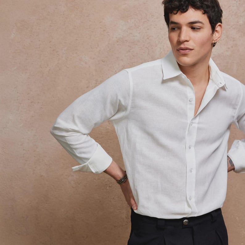 May include: A white long-sleeve shirt with a classic collar and button-down front. The shirt is made of a textured fabric and is paired with black pants. The sleeves are cuffed at the wrists.