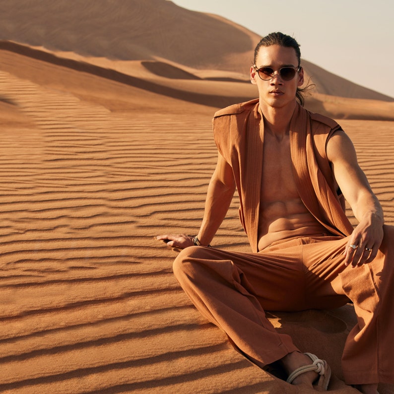 May include: A person wearing a brown sleeveless vest, matching pants, and sunglasses sits in the desert. The outfit is a solid color. The background is a desert landscape with sand dunes.