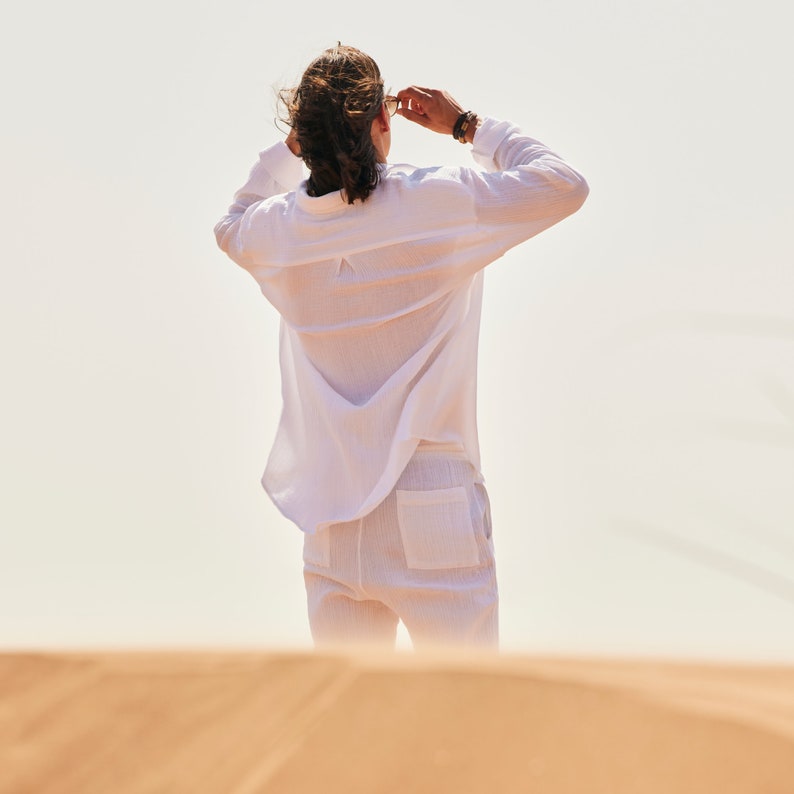 Peut inclure: Une personne porte une chemise blanche &agrave; manches longues et un pantalon blanc. La personne tient des lunettes de soleil et regarde vers le haut. Les v&ecirc;tements semblent &ecirc;tre faits d'un tissu l&eacute;ger, adapt&eacute; aux climats chauds.