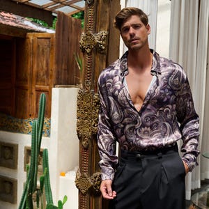 May include: A long-sleeved paisley print shirt in shades of purple, brown, and cream. The shirt is paired with black pleated trousers. The shirt has a button-down collar and is unbuttoned at the chest.
