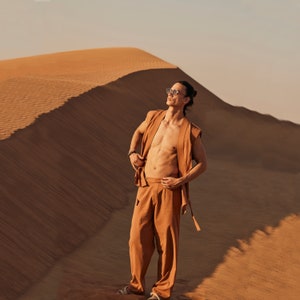 May include: A person wearing a sleeveless, open-front, rust-colored vest and matching pants stands in a desert landscape. The outfit is paired with sunglasses. The background features a large sand dune under a clear sky.