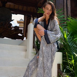 May include: A long, flowing robe with a snakeskin pattern in shades of gray and white, accented with navy blue trim. The robe is worn outdoors, draped over a white railing, suggesting resort wear or loungewear.