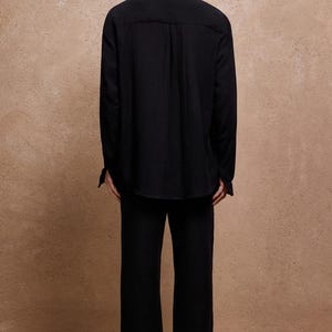 May include: A person wearing a black long-sleeve shirt and matching pants. The shirt has a collar and buttoned cuffs. The pants are wide-legged. The background is a solid, textured, light brown color.
