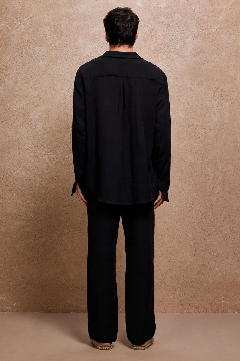 May include: A person wearing a black long-sleeve shirt and matching pants. The shirt has a collar and buttoned cuffs. The pants are wide-legged. The background is a solid, textured, light brown color.