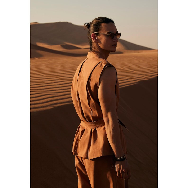 May include: A person wearing a tan vest and trousers, with a belt. The person is wearing sunglasses and a bracelet. The background is a desert landscape with sand dunes.
