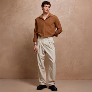 May include: A man wearing a brown long-sleeve shirt and cream-colored pleated trousers. The shirt is buttoned and has a collar. The trousers are high-waisted and have a wide leg. The man is also wearing black shoes.