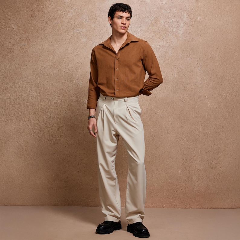 May include: A man wearing a brown long-sleeve shirt and cream-colored pleated trousers. The shirt is buttoned and has a collar. The trousers are high-waisted and have a wide leg. The man is also wearing black shoes.