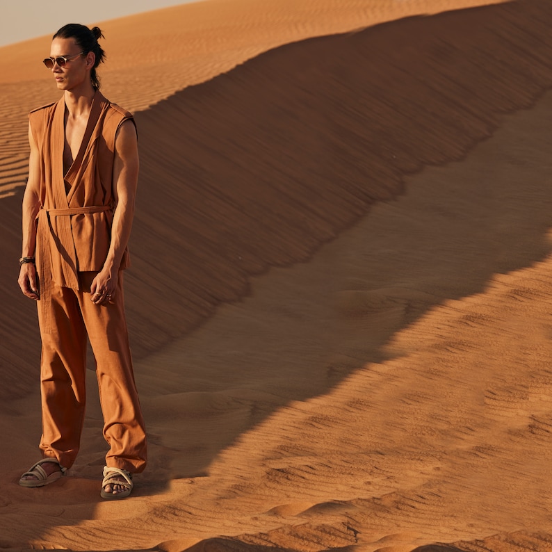 May include: A person wearing a tan sleeveless top, matching pants, and sandals stands on a sand dune. The outfit has a wrap-style top with a belt. The person is wearing sunglasses and has their hair pulled back.
