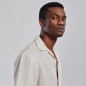 May include: A man wearing a cream-colored linen shirt with thin black vertical stripes. The shirt has a collar and a button-up front.
