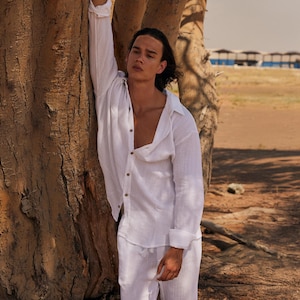 May include: A white linen shirt and matching trousers. The long-sleeved shirt is unbuttoned at the chest. The model is leaning against a tree trunk. The background is a desert landscape with buildings.