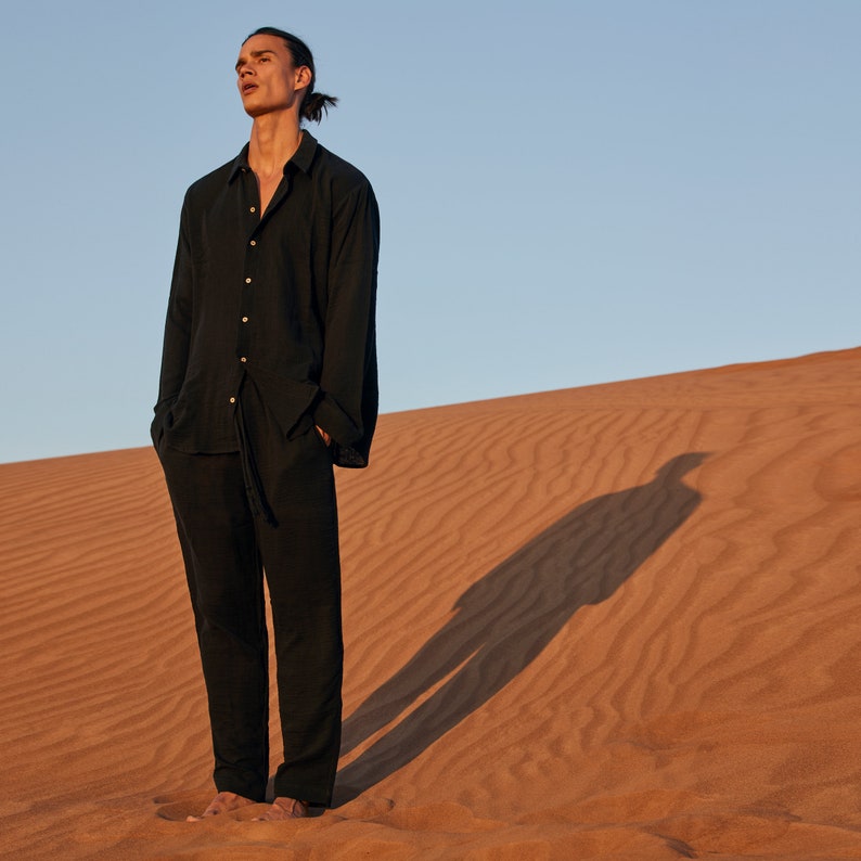 May include: Black linen shirt and pants set. The long-sleeved shirt has a button-down front and the pants have a drawstring waist. The outfit is worn in a desert setting with a blue sky background.