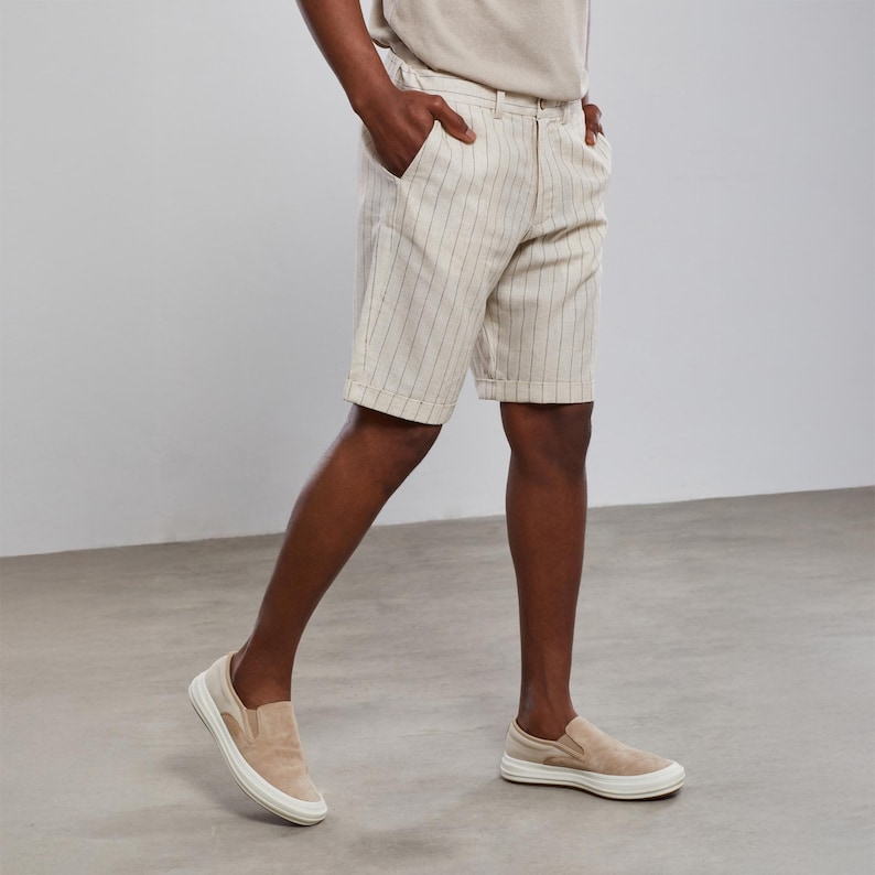 May include: A pair of beige linen shorts with thin vertical stripes. The shorts have a button and zipper closure and belt loops. The model is wearing the shorts with a beige t-shirt and beige slip-on shoes.