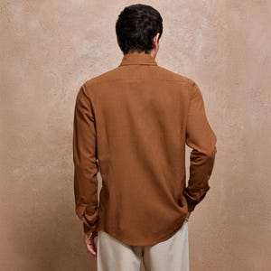 May include: A long-sleeved, button-down shirt in a warm brown hue. The shirt features a classic collar and buttoned cuffs. The model is wearing beige pants. The background is a textured, neutral color.