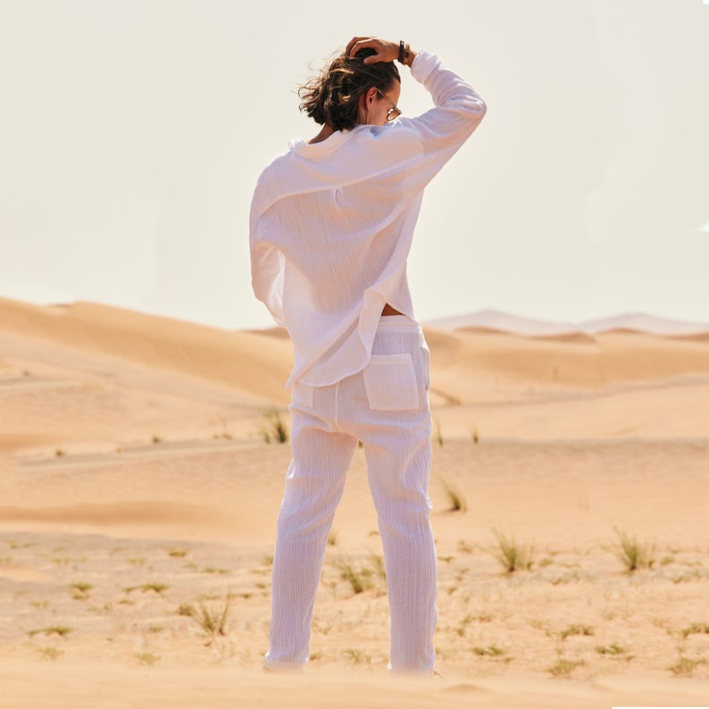 Peut inclure: Une personne porte une chemise blanche &agrave; manches longues et un pantalon blanc dans un paysage d&eacute;sertique. La chemise est ample et le pantalon a des poches. L'arri&egrave;re-plan pr&eacute;sente des dunes de sable et un ciel clair.