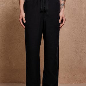 May include: Black drawstring pants with a relaxed fit. The pants have a visible drawstring at the waist and a loose, straight leg. The fabric appears to have a subtle texture. The pants are paired with light brown suede shoes.