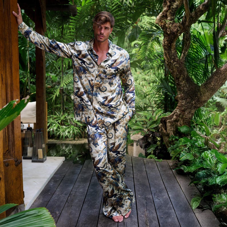 Puede incluir: Un conjunto de pijama a juego con una camisa con botones y pantalones. El conjunto es de color crema con un patr&oacute;n repetido de p&aacute;jaros estilizados azules y marrones y elementos florales. La camisa de manga larga y los pantalones son de material sedoso.