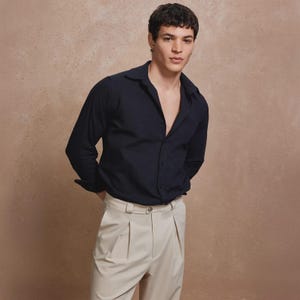 May include: A navy blue button-down shirt with long sleeves and a spread collar is paired with light beige pleated trousers. The shirt is unbuttoned at the top. The model is standing with their hands behind their back.