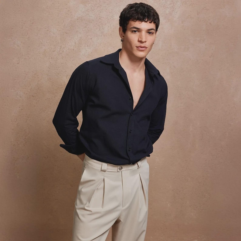 May include: A navy blue button-down shirt with long sleeves and a spread collar is paired with light beige pleated trousers. The shirt is unbuttoned at the top. The model is standing with their hands behind their back.
