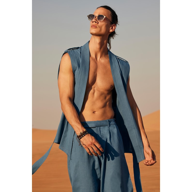 May include: A person wearing a sleeveless blue vest and matching trousers, with sunglasses, in a desert setting. The vest has a draped design and the trousers are high-waisted. The person is also wearing a bracelet.