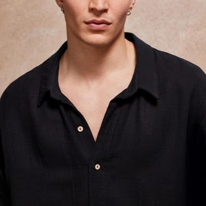 May include: Black button-down shirt with a classic collar. The shirt has a relaxed fit and is made of a lightweight fabric. The buttons are a light beige color. The shirt is a solid black color.