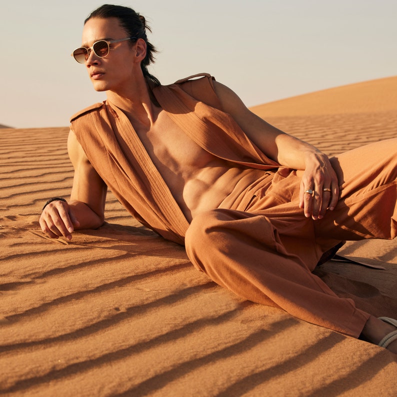 May include: A person wearing a tan sleeveless vest and matching pants, lying on sand dunes. The person is also wearing sunglasses and rings. The outfit is a solid color, and the background is a desert landscape.