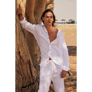Peut inclure: Ensemble chemise et pantalon en lin blanc. La chemise &agrave; manches longues a une fermeture boutonn&eacute;e sur le devant et une coupe d&eacute;contract&eacute;e. Le pantalon a une taille &agrave; cordon. L'ensemble est port&eacute; par une personne appuy&eacute;e contre un arbre.