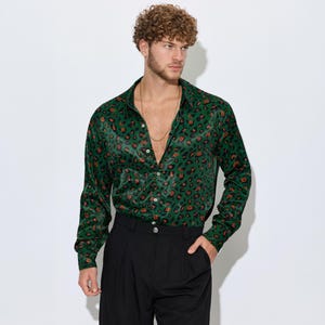 May include: A long-sleeved green button-down shirt with a leopard print pattern in orange and black. The shirt is paired with black pleated trousers. The model is wearing a gold chain.