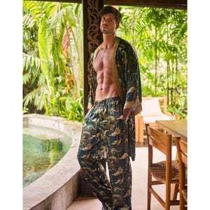May include: A teal and gold patterned pajama set, including pants and a robe with gold trim. The pajama set features a dragon and cloud design. The person is standing near a pool and wooden table.