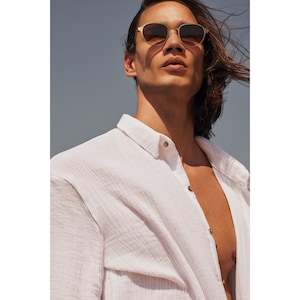 Peut inclure: Lunettes de soleil &agrave; monture dor&eacute;e et verres bruns. La personne porte des lunettes de soleil et une chemise blanche boutonn&eacute;e. La chemise est d&eacute;boutonn&eacute;e au niveau de la poitrine. L'arri&egrave;re-plan est bleu clair.
