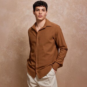 May include: A brown long-sleeve button-down shirt with a classic collar. The shirt is paired with off-white pants. The model is standing in front of a neutral-toned background.