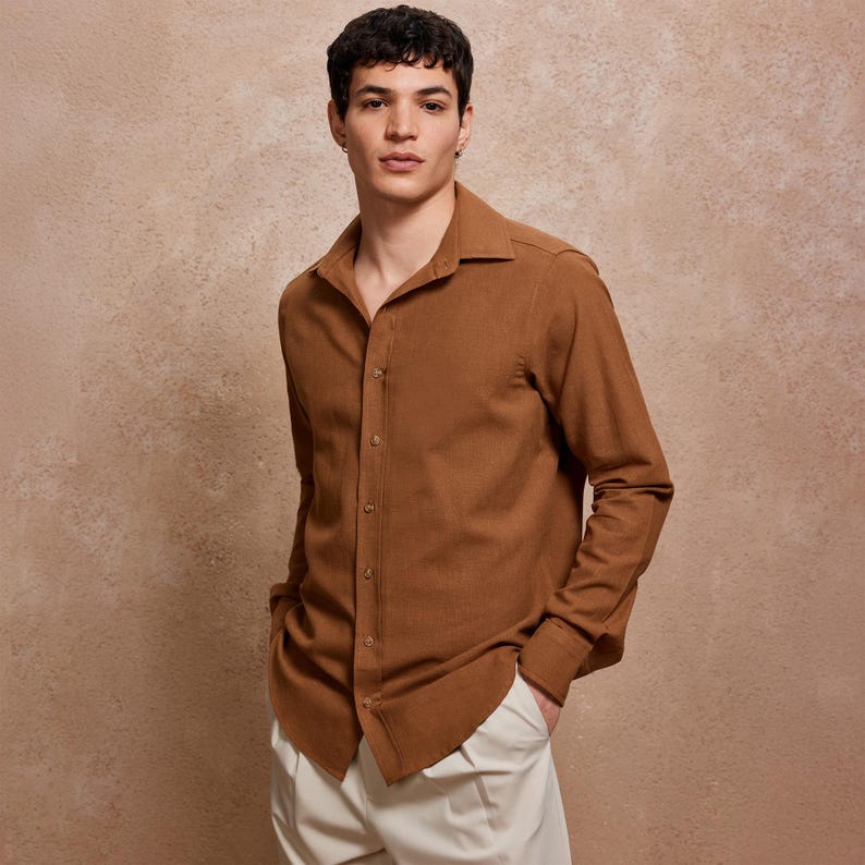 May include: A brown long-sleeve button-down shirt with a classic collar. The shirt is paired with off-white pants. The model is standing in front of a neutral-toned background.