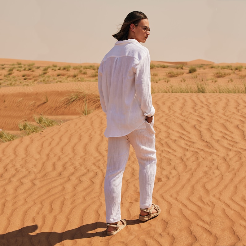 Peut inclure: Un ensemble chemise et pantalon en lin blanc. La chemise &agrave; manches longues a un devant boutonn&eacute; et le pantalon a des poches. La tenue est associ&eacute;e &agrave; des sandales. La personne se tient dans un paysage d&eacute;sertique avec des dunes de sable.