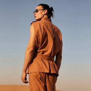 May include: A person wearing a rust-colored sleeveless top and matching pants, with a belted waist. The outfit is made of a textured fabric. The person is wearing sunglasses and has their hair pulled back. The background is a desert landscape with a blue sky.