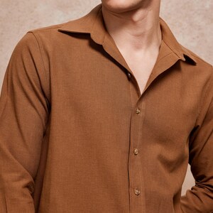 May include: A long-sleeved, button-down shirt in a warm brown hue. The shirt features a camp collar and is buttoned up the front with matching brown buttons. The fabric appears to be a textured linen or similar material.