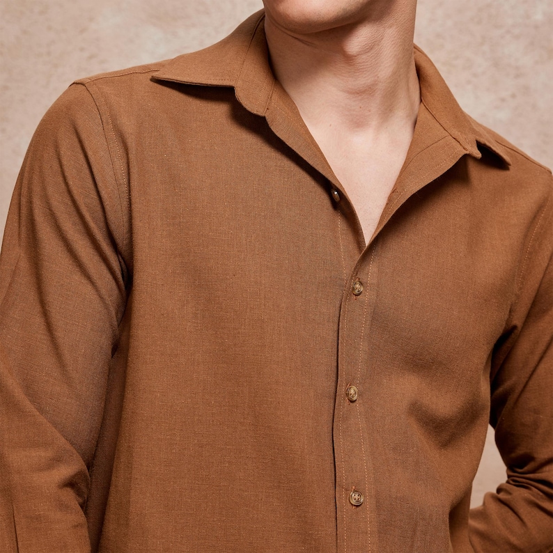 May include: A long-sleeved, button-down shirt in a warm brown hue. The shirt features a camp collar and is buttoned up the front with matching brown buttons. The fabric appears to be a textured linen or similar material.