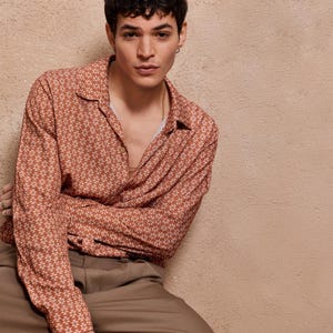 May include: A man wearing a long-sleeved shirt with a brown and white geometric pattern. The shirt has a collared neckline and a button-up front. The man is wearing brown pants.