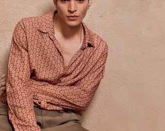 May include: A man wearing a long-sleeved shirt with a brown and white geometric pattern. The shirt has a collared neckline and a button-up front. The man is wearing brown pants.