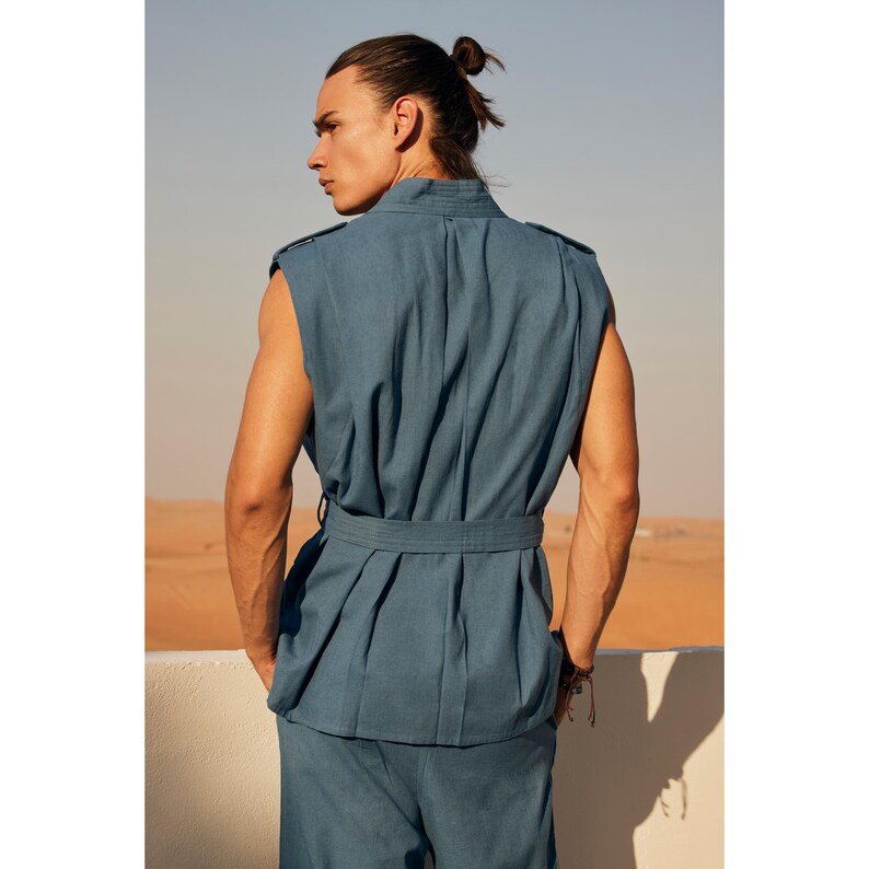 May include: A blue sleeveless vest and matching trousers. The vest features a belted waist and shoulder epaulettes. The model is standing outdoors with a desert landscape in the background. The outfit is made of a textured fabric.