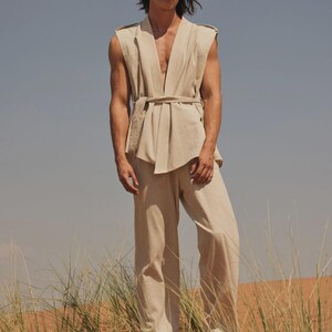 May include: A beige linen outfit. The ensemble includes a sleeveless top with a belt and wide-leg trousers. The model is standing in a desert landscape with tall grass and a blue sky. The outfit is a neutral colour.