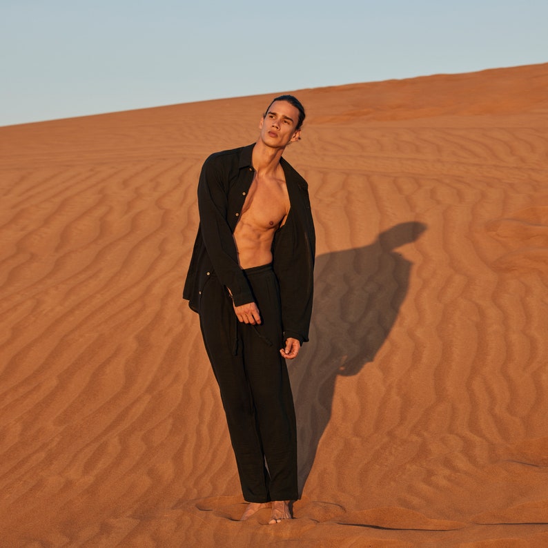 May include: A person wearing a black button-down shirt and black pants stands in the desert. The shirt is unbuttoned, revealing the person's torso. The background is a sand dune under a clear sky. The person is barefoot.
