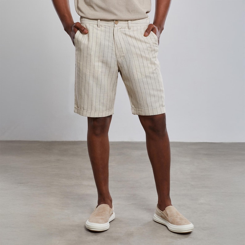 May include: A pair of beige linen shorts with thin vertical white stripes. The shorts have a button and zipper closure and two front pockets.