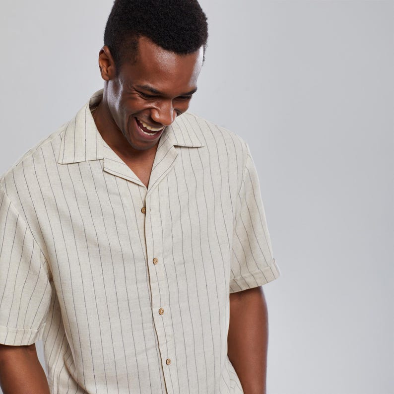 May include: A man wearing a white and black striped short-sleeved shirt with a collared neckline. He is smiling and looking down.
