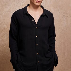 May include: A black button-down shirt with long sleeves. The shirt has a classic collar and is buttoned up the front with several buttons. The fabric appears to be a lightweight material. The model is wearing the shirt with black pants.