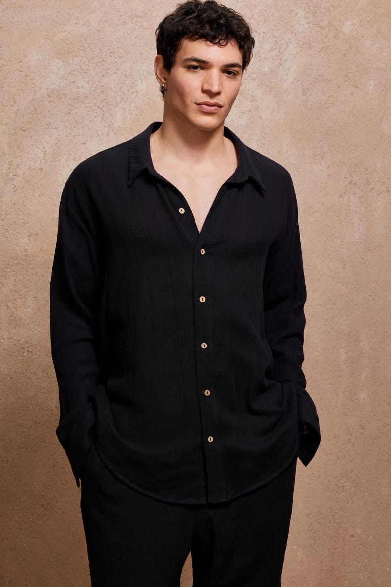 May include: A black button-down shirt with long sleeves. The shirt has a classic collar and is buttoned up the front with several buttons. The fabric appears to be a lightweight material. The model is wearing the shirt with black pants.