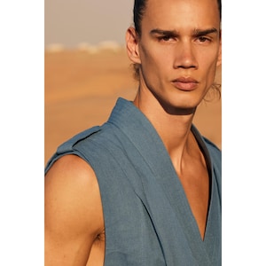 May include: A sleeveless, blue-grey top with a V-neck and structured shoulders. The fabric appears to be linen or a similar lightweight material. The background is a blurred desert landscape.
