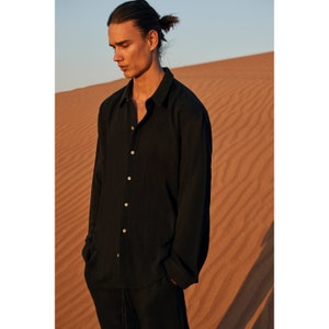 May include: Black button-down shirt with long sleeves and a relaxed fit. The shirt has a classic collar and is paired with black pants. The model is standing in a desert landscape with sand dunes in the background.