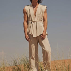 May include: A beige linen outfit consisting of a sleeveless top with a belt and wide-leg pants. The model is standing in a desert landscape with tall grass and a blue sky. The outfit is a neutral color.