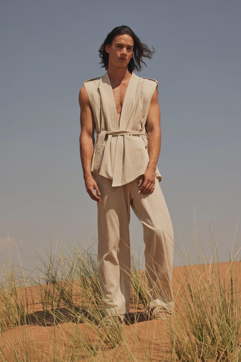 May include: A beige linen outfit consisting of a sleeveless top with a belt and wide-leg pants. The model is standing in a desert landscape with tall grass and a blue sky. The outfit is a neutral color.