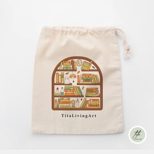May include: A beige drawstring bag featuring a brown arched bookshelf design filled with books, a ghost, and potion bottles. The text "TitaLivingArt" is printed below the bookshelf illustration. A small logo is in the bottom right corner.