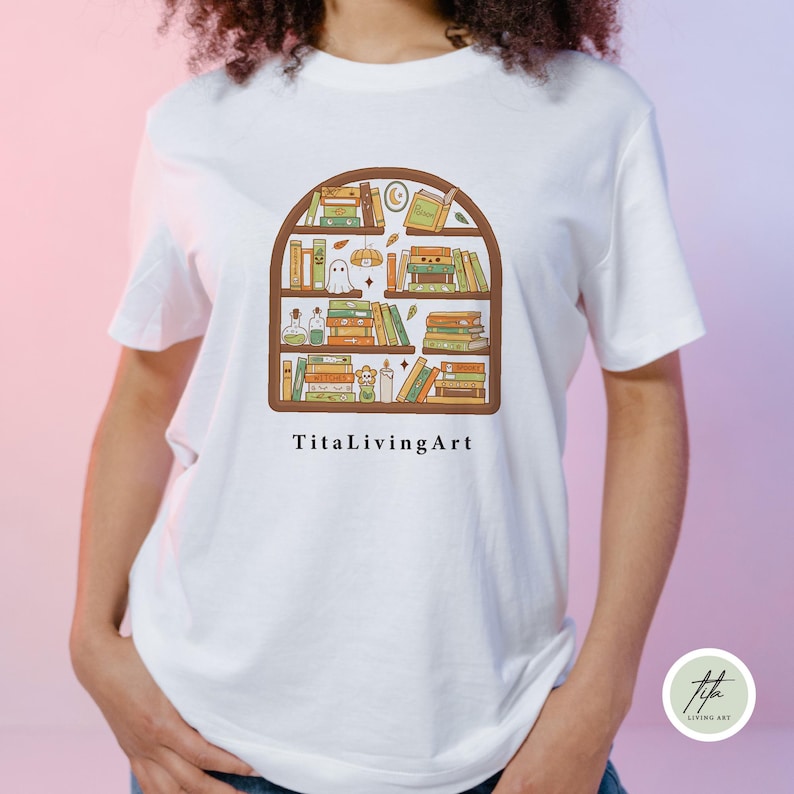 May include: White t-shirt featuring a whimsical illustration of a bookshelf filled with books, a ghost, and other decorative items. The design is in shades of brown, green, and orange. The text "TitaLivingArt" is printed below the bookshelf.